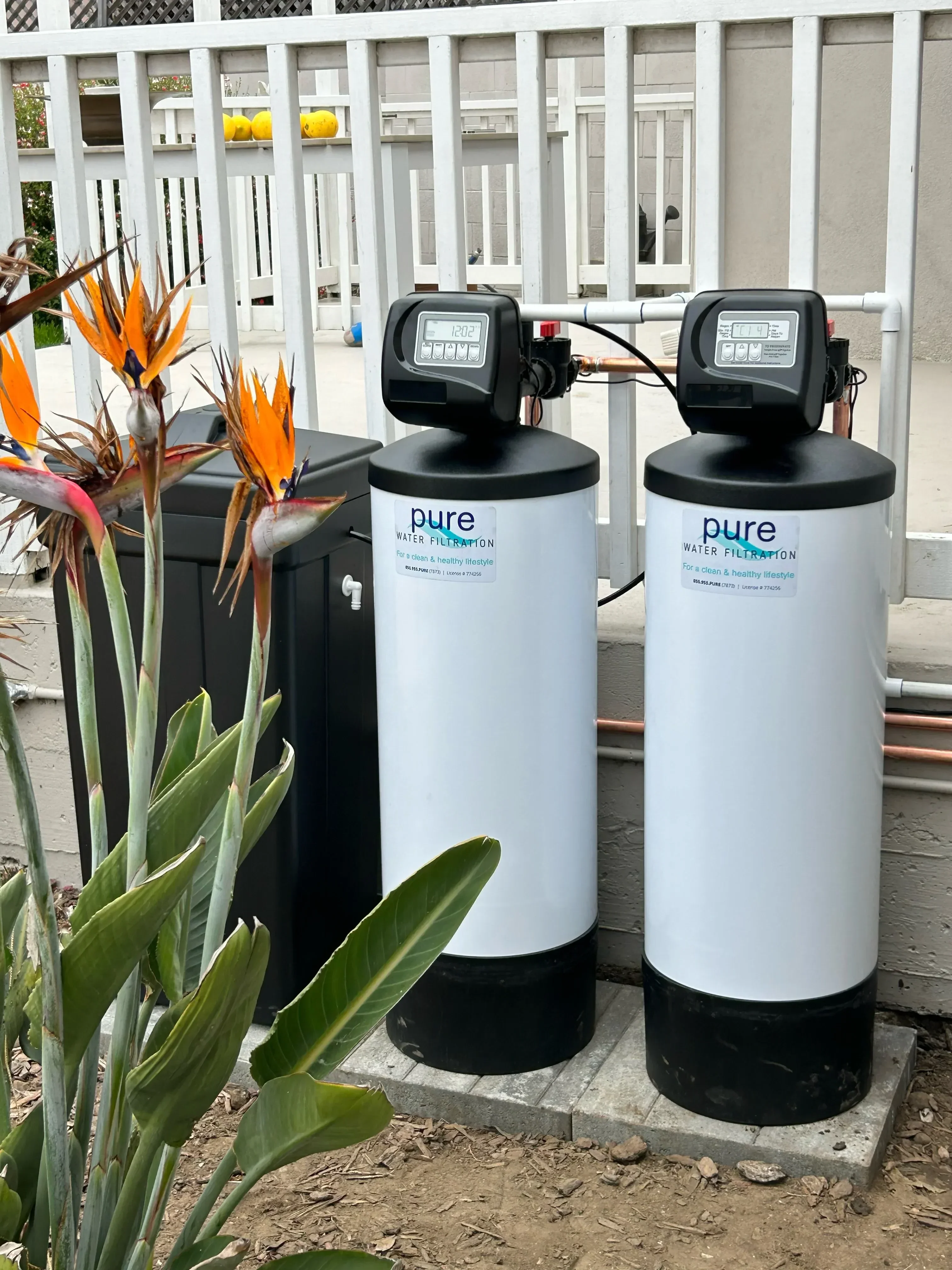 Two white water softener tanks with black control valves mounted on white rack with copper piping