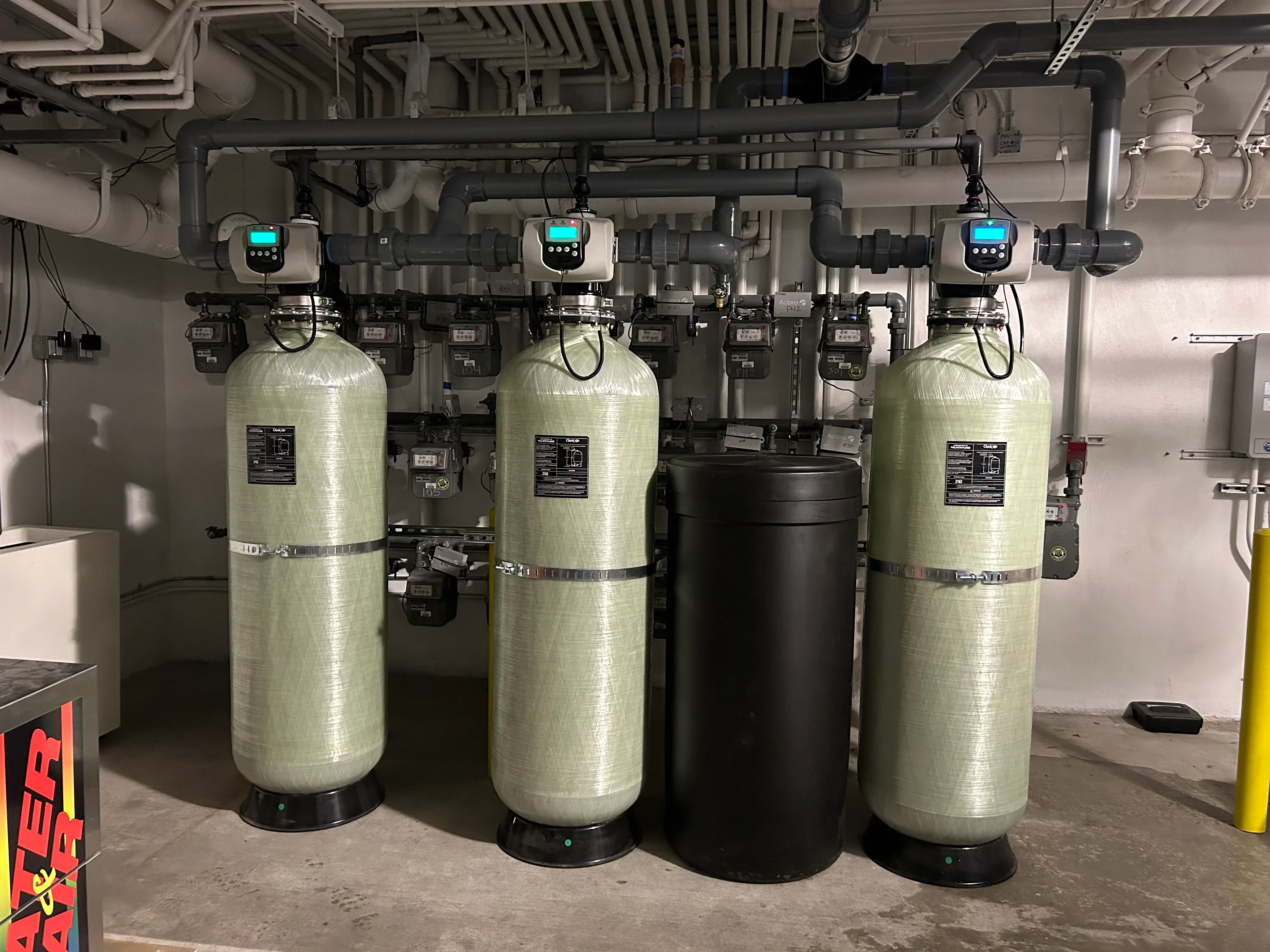 Three fiberglass water softening tanks with digital meters and black carbon filter vessel, overhead PVC piping system
